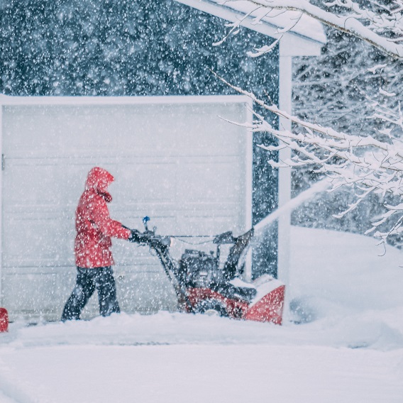 Person kör snömaskin utanför garage i snölandskap