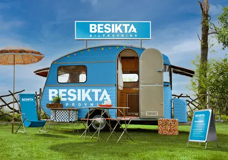 Ljusblå husvagn med Besikta-loggan på campingplats, gräs och blå himmel